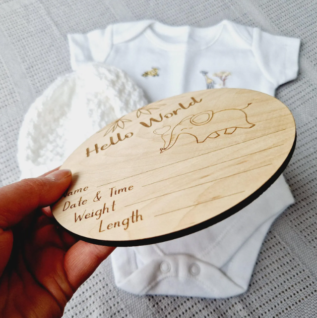 A wooden baby announcement card is being held up, with a white onesie and a white hat visible in the background.