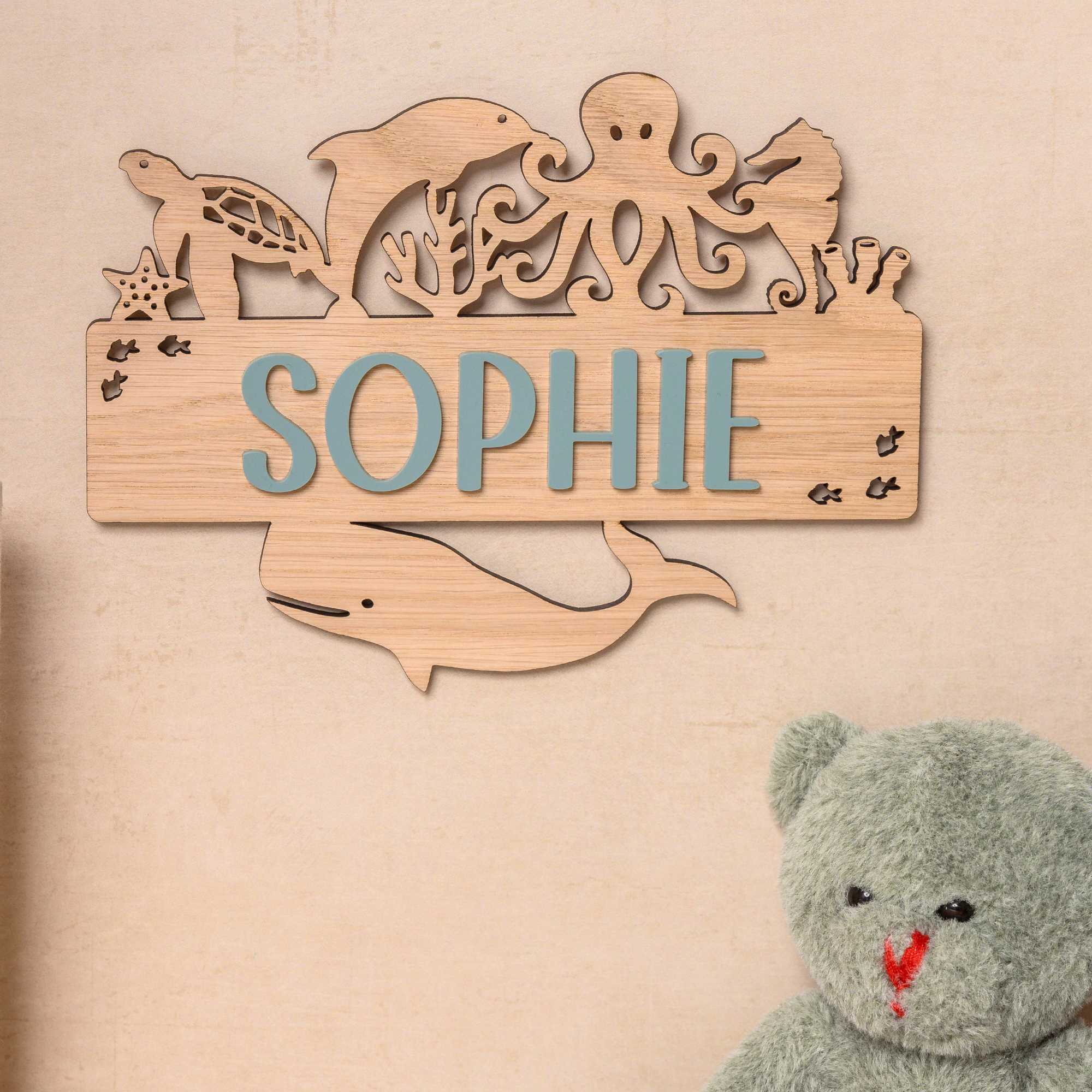 A wooden nameplate with the name "Sophie" is hanging on a wall, surrounded by various sea animal silhouettes. Next to the nameplate is a stuffed teddy bear.