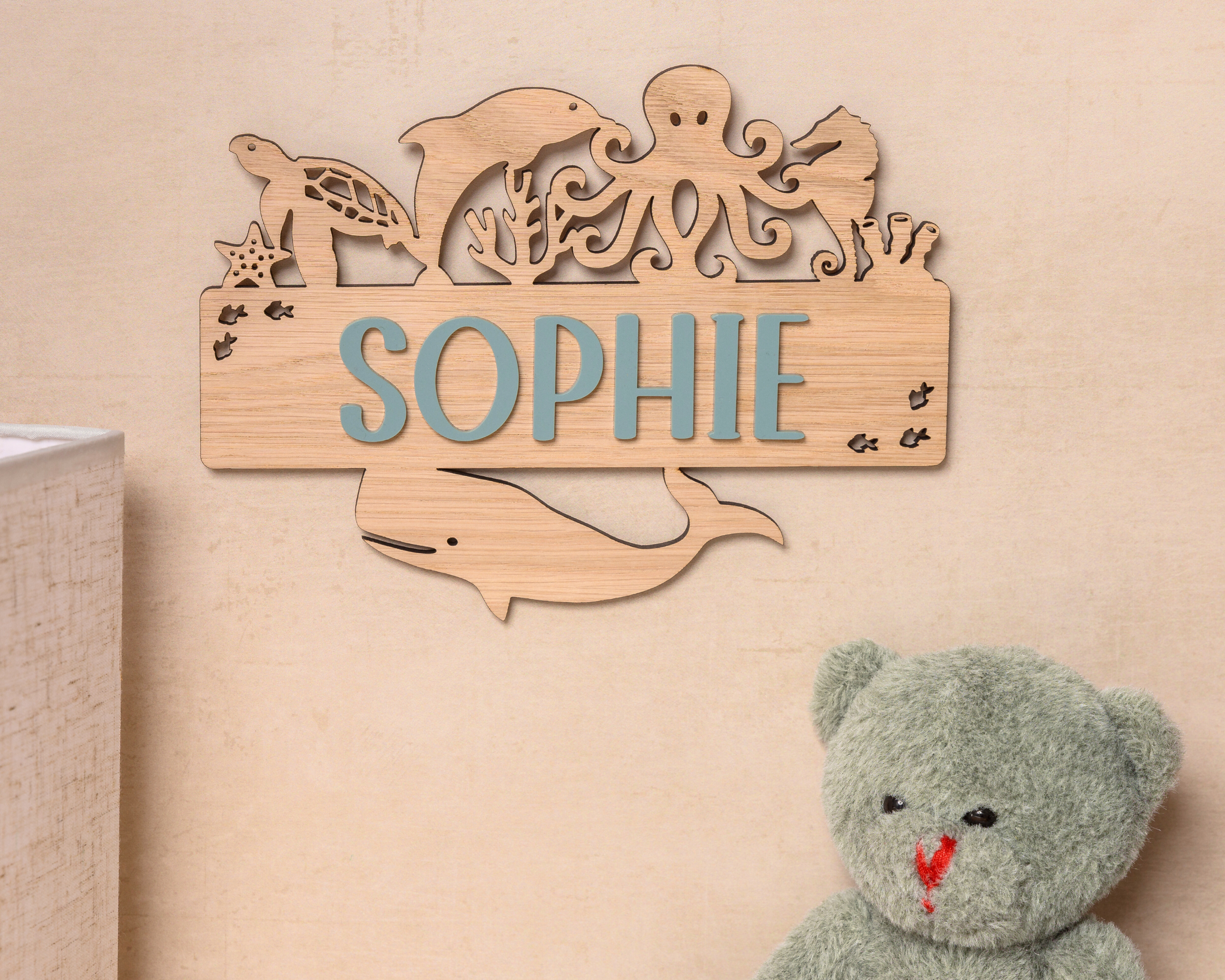 A wooden nameplate with the name "Sophie" is hanging on a wall, surrounded by various sea animal silhouettes. Next to the nameplate is a stuffed teddy bear.