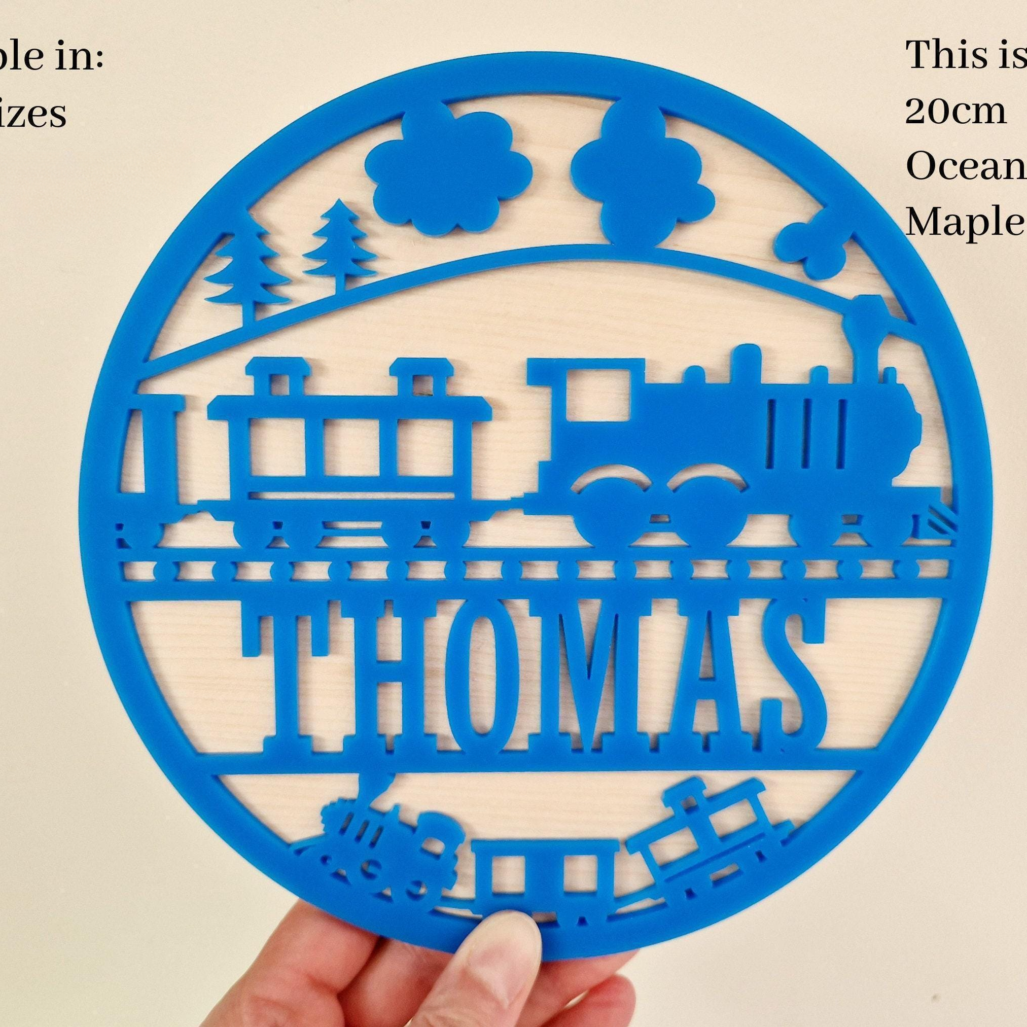 A hand holding a blue circular cutout with a train design and the name "THOMAS" written on it.