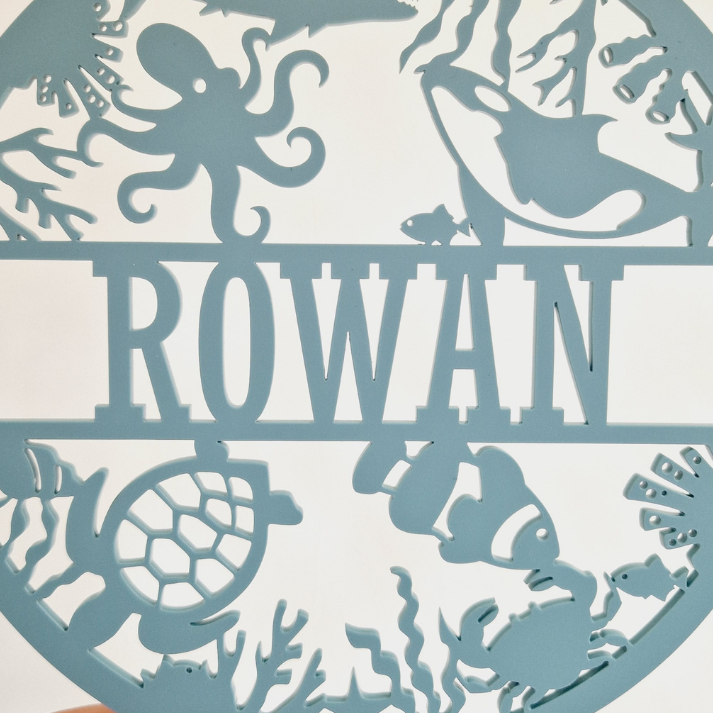 a round sign with the name "Rowan" on it, available in 25+ colors and 3 sizes. The sign is described as a thoughtful gift for various occasions, such as birthdays, Christmas, baby showers, and other special events. It is also wrapped in tissue paper, ready for gifting.