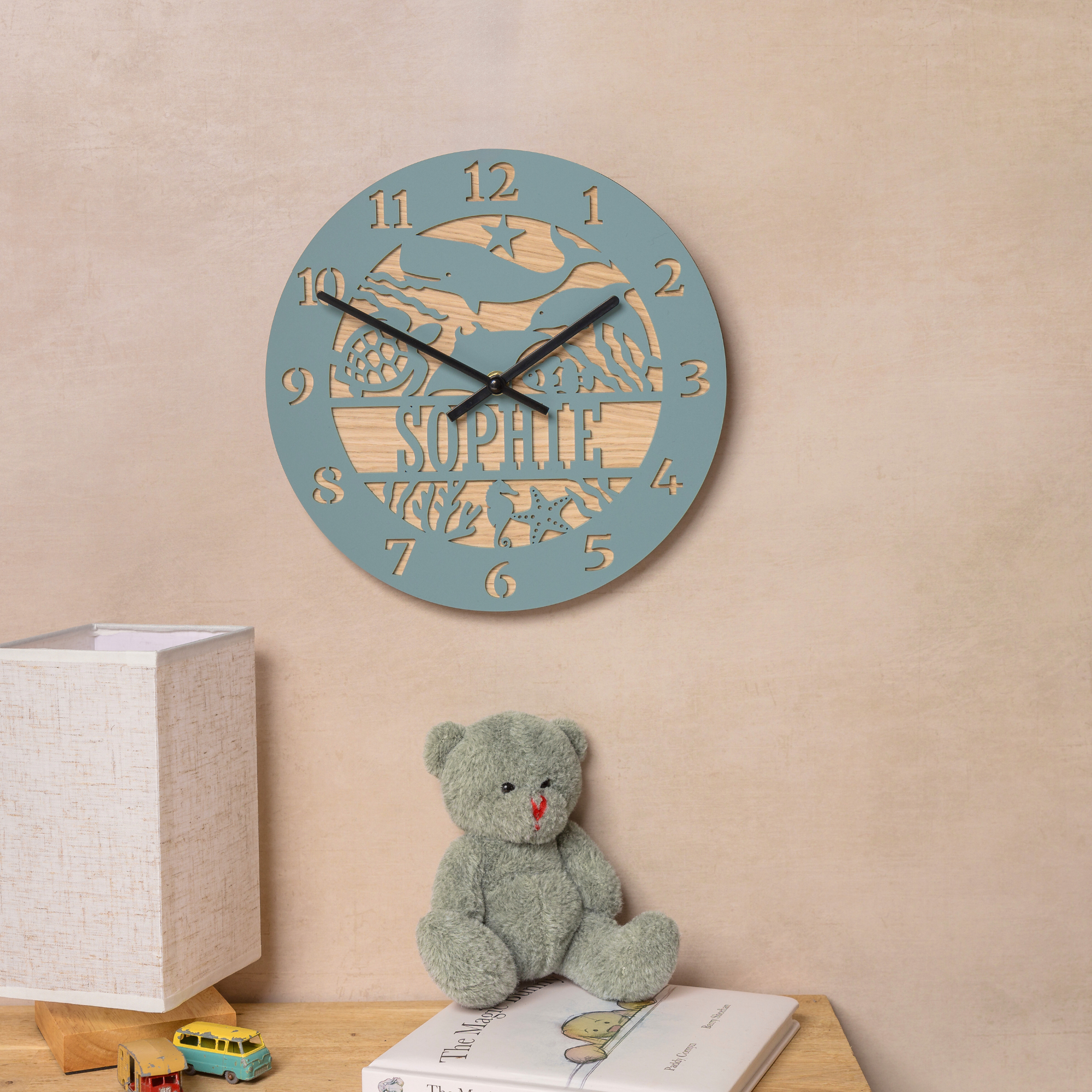 a wall clock with a personalized name, a teddy bear, and a lamp on a wooden table.