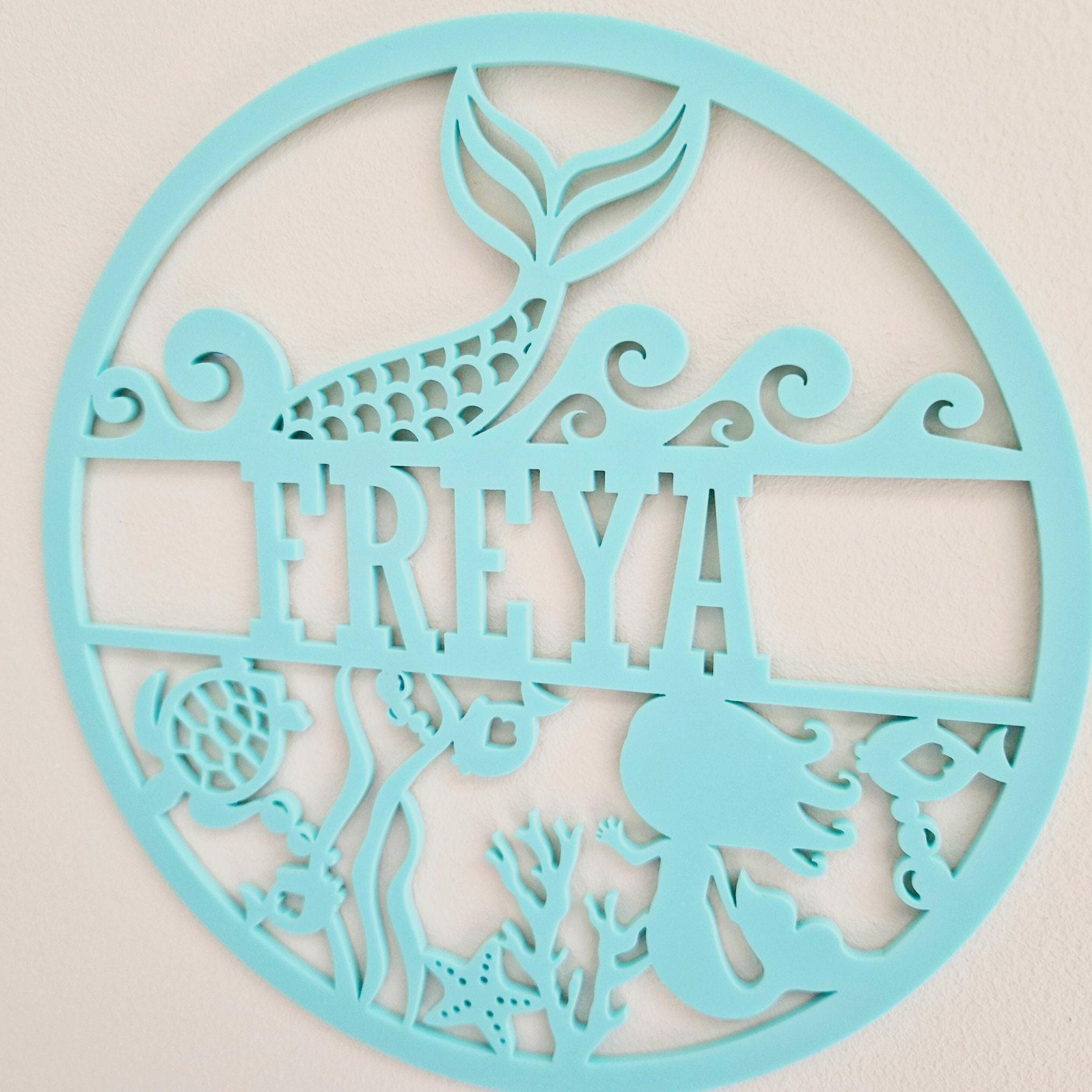A round, light blue wall hanging featuring a mermaid silhouette and various marine-themed elements, including a fish, sea star, and coral.