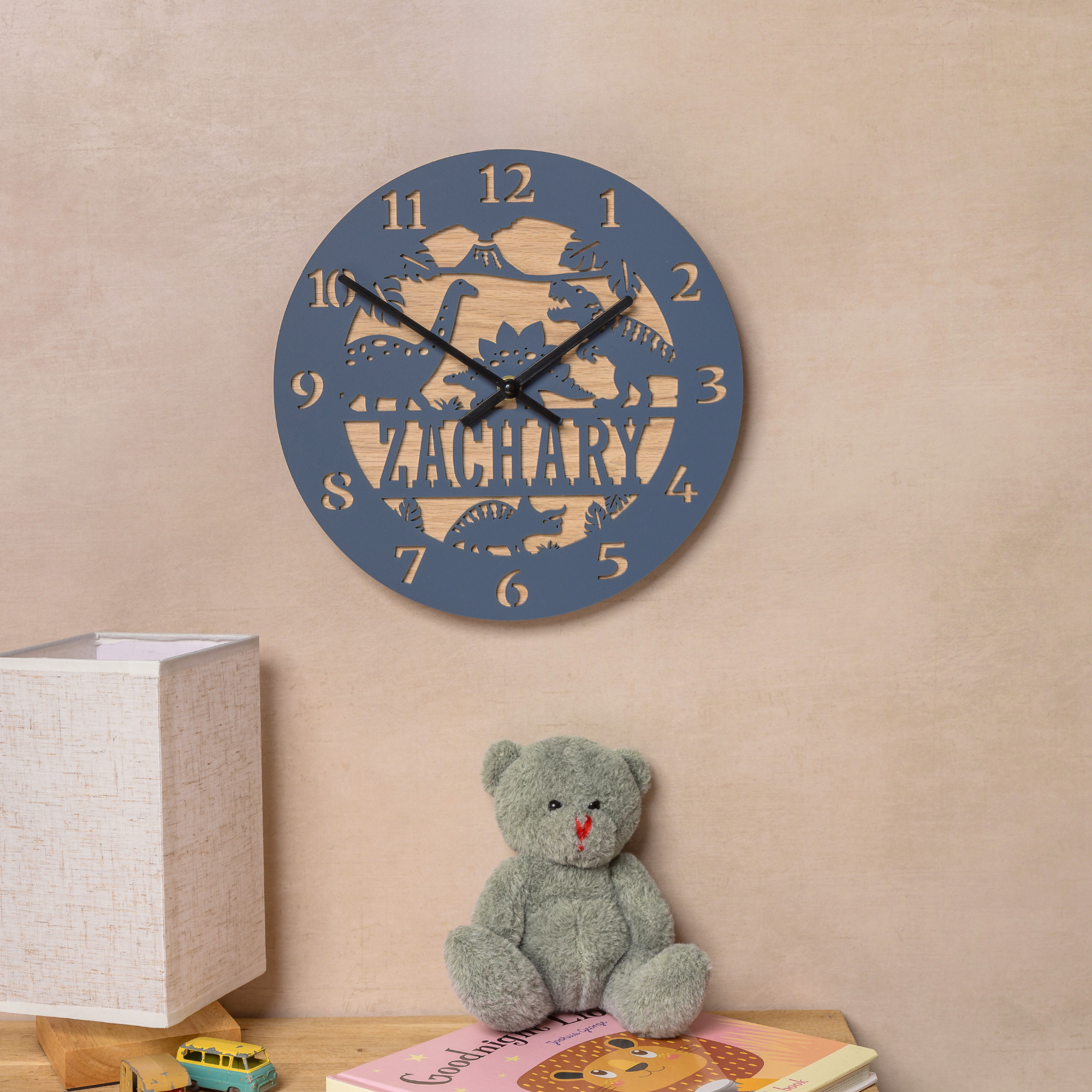 A wall clock with the name "ZACHARY" printed on it, along with a teddy bear and a children's book on a shelf.