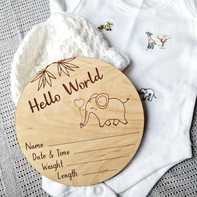 A wooden baby announcement card with a name and date and time information, along with a baby's weight and length, is placed next to a white onesie with animal prints.