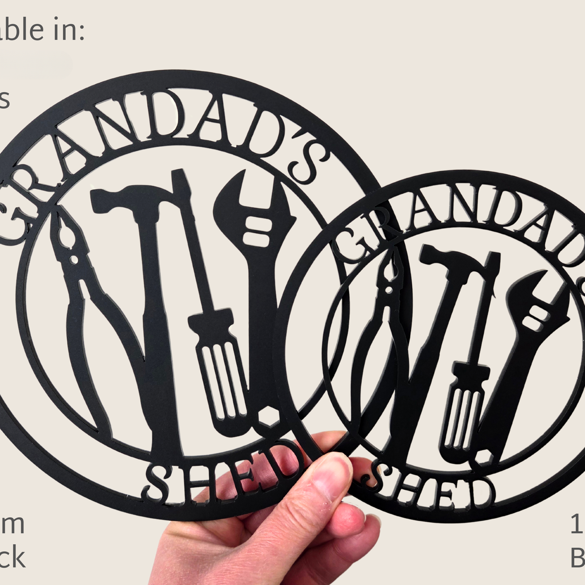 A person is holding two metal cutouts of a design featuring tools and the text "GRANDDAD'S SHED".