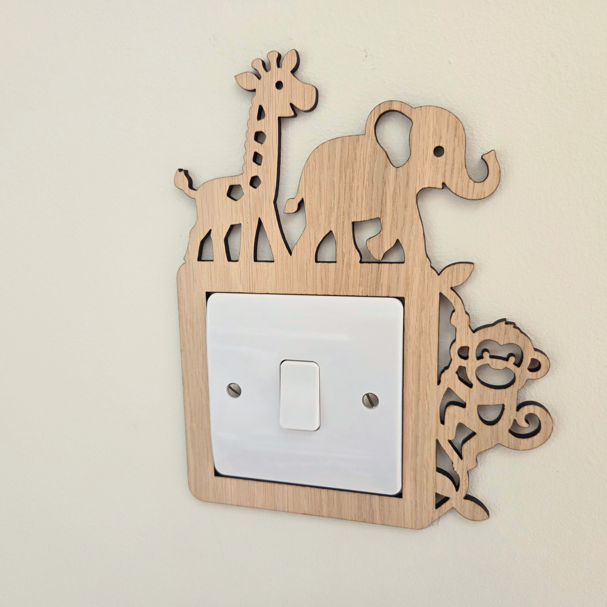A light natural wood animal silhouette wall decor piece mounted on a beige wall, featuring a giraffe, elephant, and monkey design around a white single-toggle light switch plate.