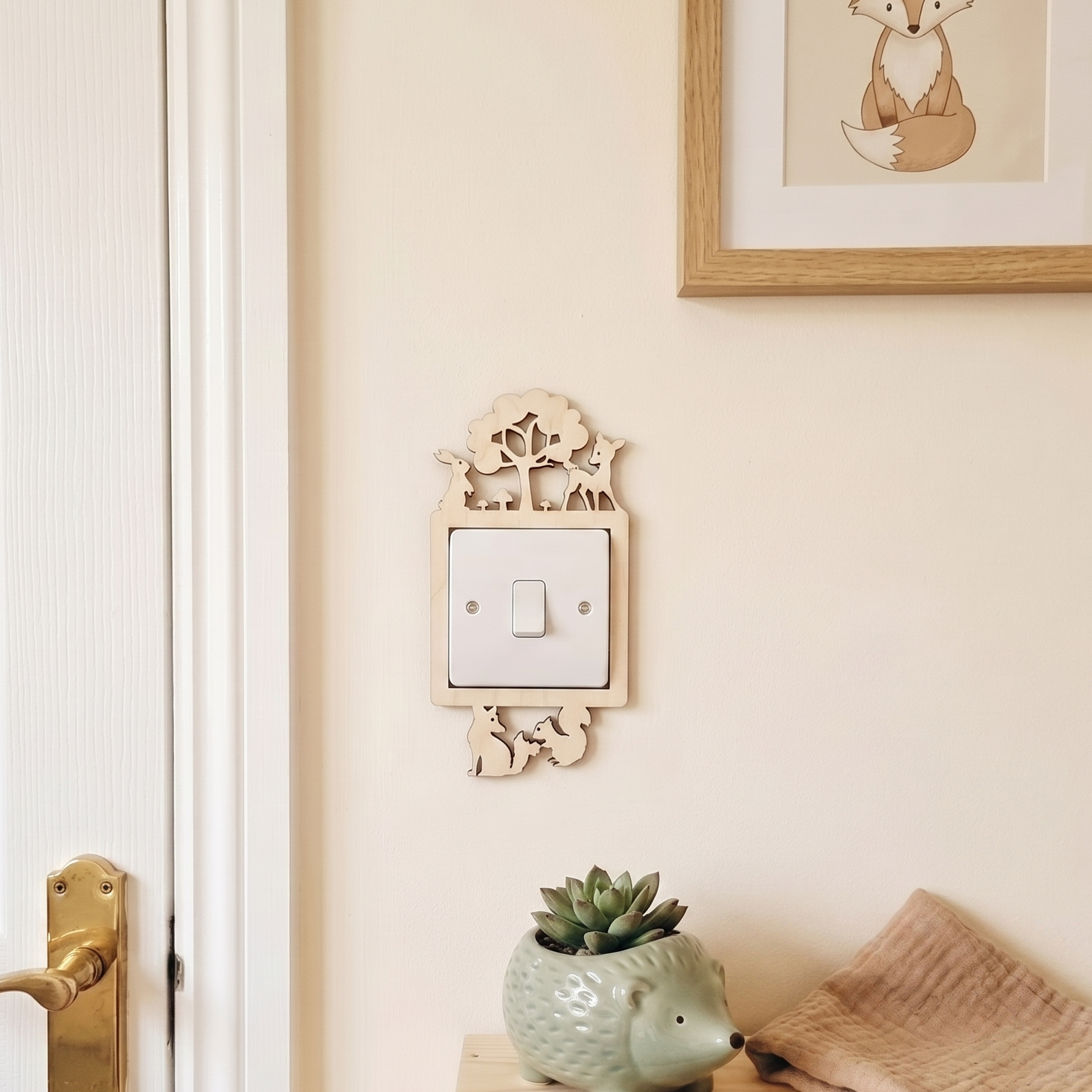 Decorative wooden wall switch plate with woodland forest animal design and underneath, a succulent plant and folded blanket on a bench.