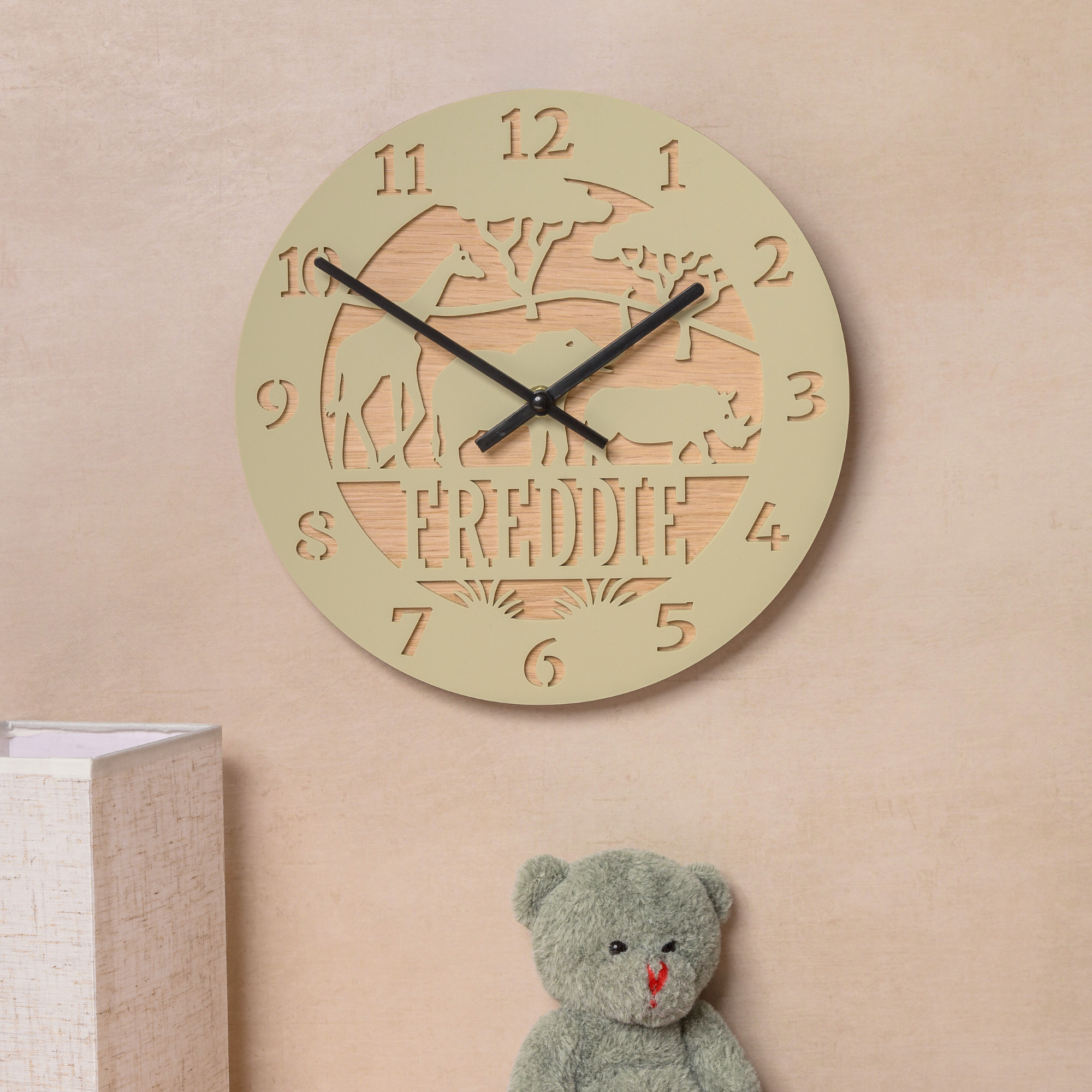 a wall clock with the name "FREDDIE" carved into it, and a small stuffed teddy bear is placed next to it on a wall.