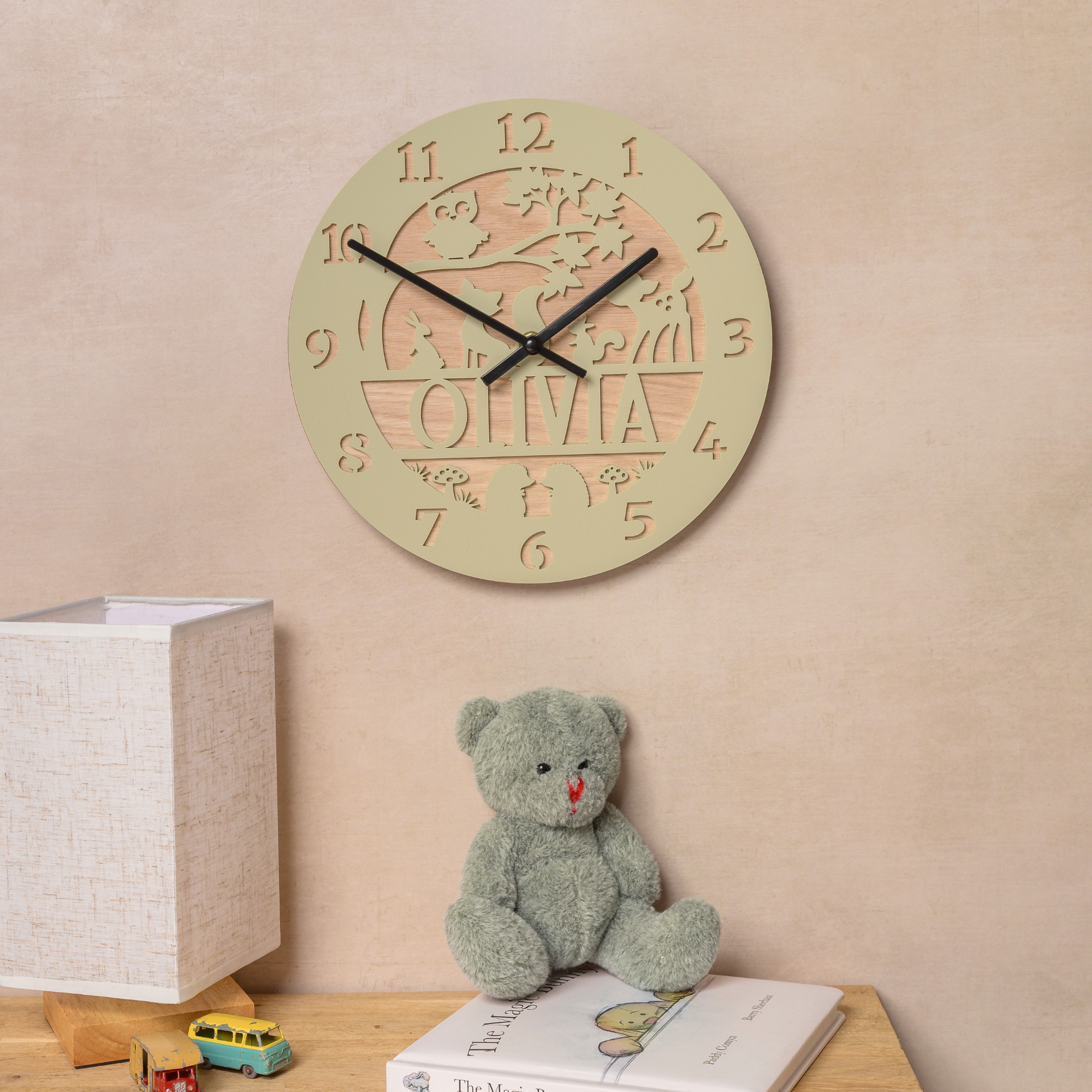 a wooden clock with the name "Olivia" carved into it, hanging on a wall above a wooden table with a teddy bear and a book.
