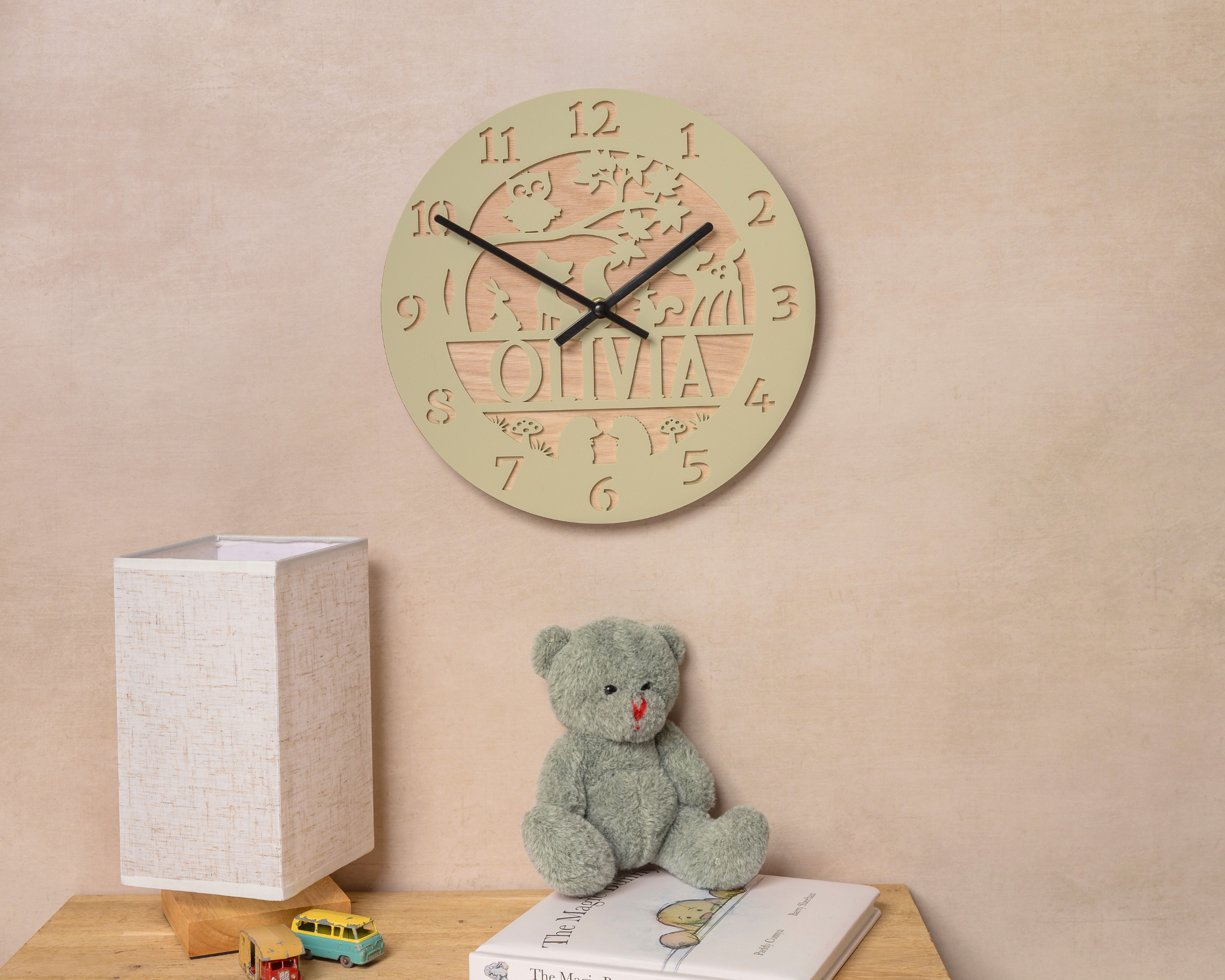 a wooden clock with the name "Olivia" carved into it, hanging on a wall above a wooden table with a teddy bear and a book.