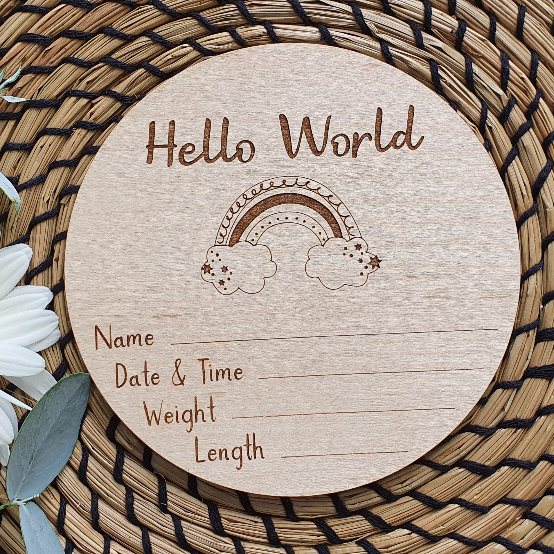 A wooden postcard with a rainbow graphic and the text "Hello World" is placed on a woven mat with a white daisy flower.