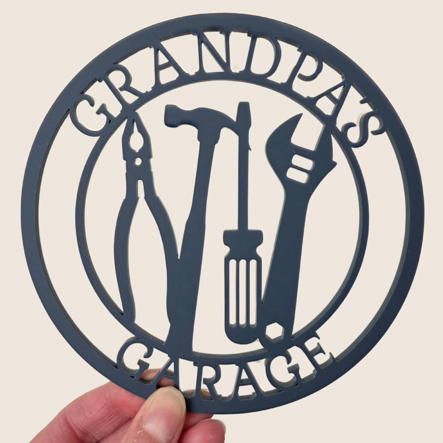 A hand holding a circular sign with the text "GRANDPA'S GARAGE" and silhouettes of various tools, such as pliers, wrenches, and screwdrivers.