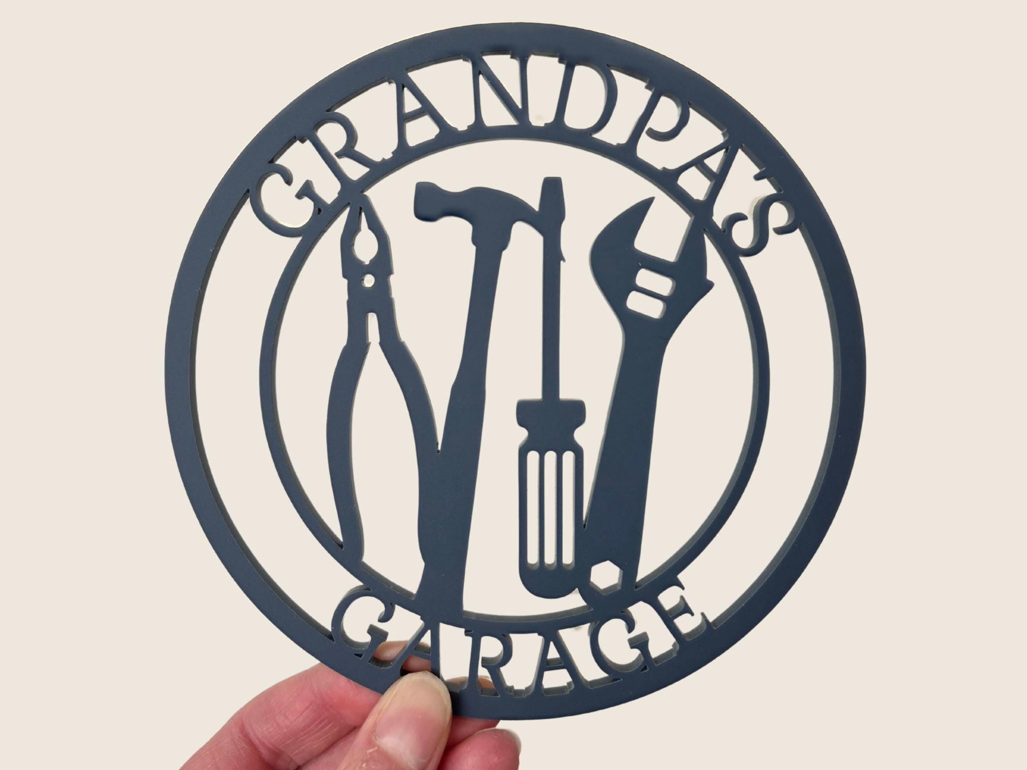 A hand holding a circular sign with the text "GRANDPA'S GARAGE" and silhouettes of various tools, such as pliers, wrenches, and screwdrivers.