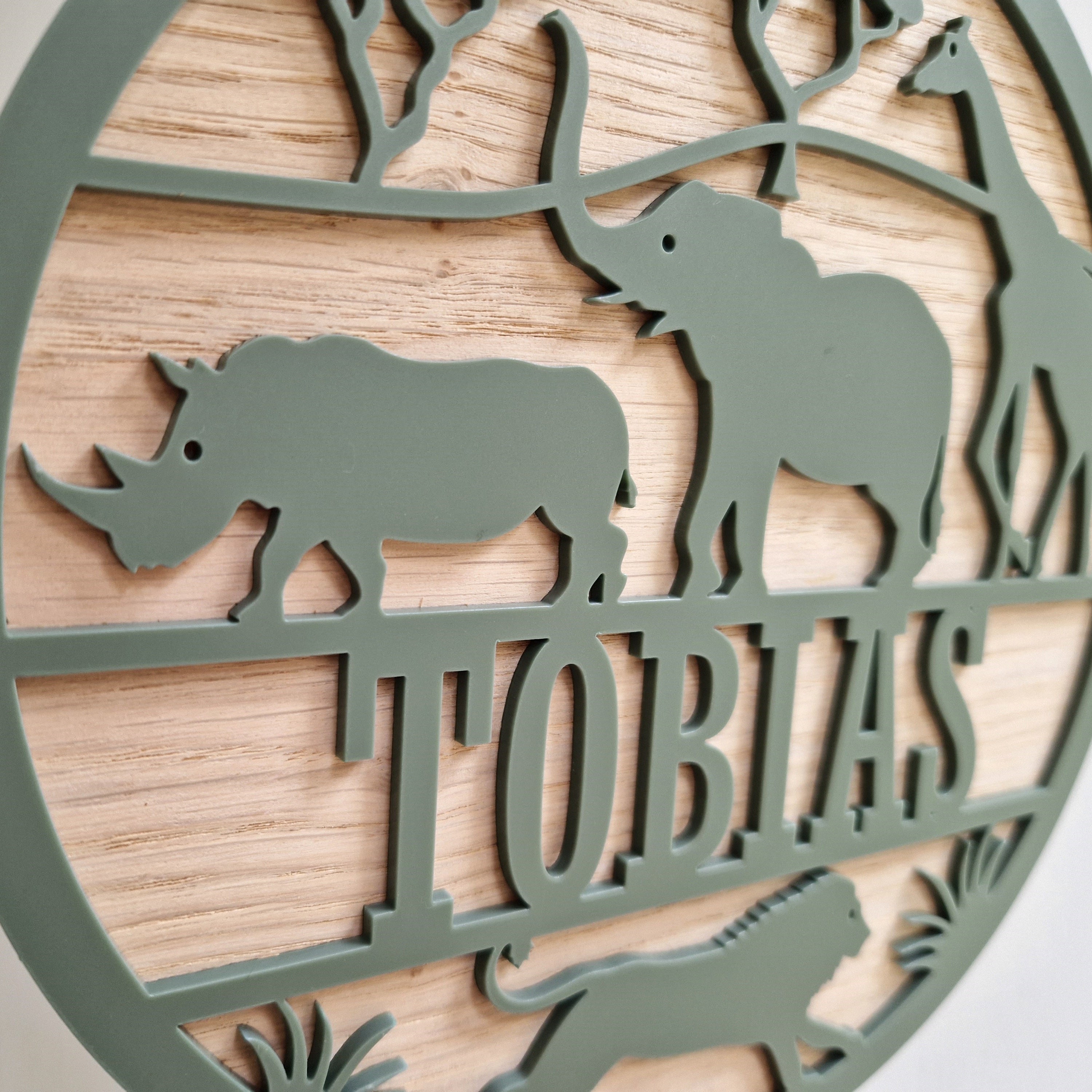 A circular metal sign with a wooden border, featuring silhouettes of a rhinoceros, an elephant, and a giraffe, along with the word "TOBIAS" written on it.
