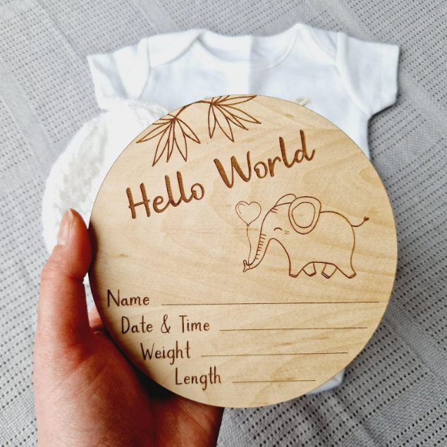 A wooden baby name card is being held up, featuring a cute elephant illustration and the text "Hello World" along with a blank space for the baby's name, date, time, weight, and length.