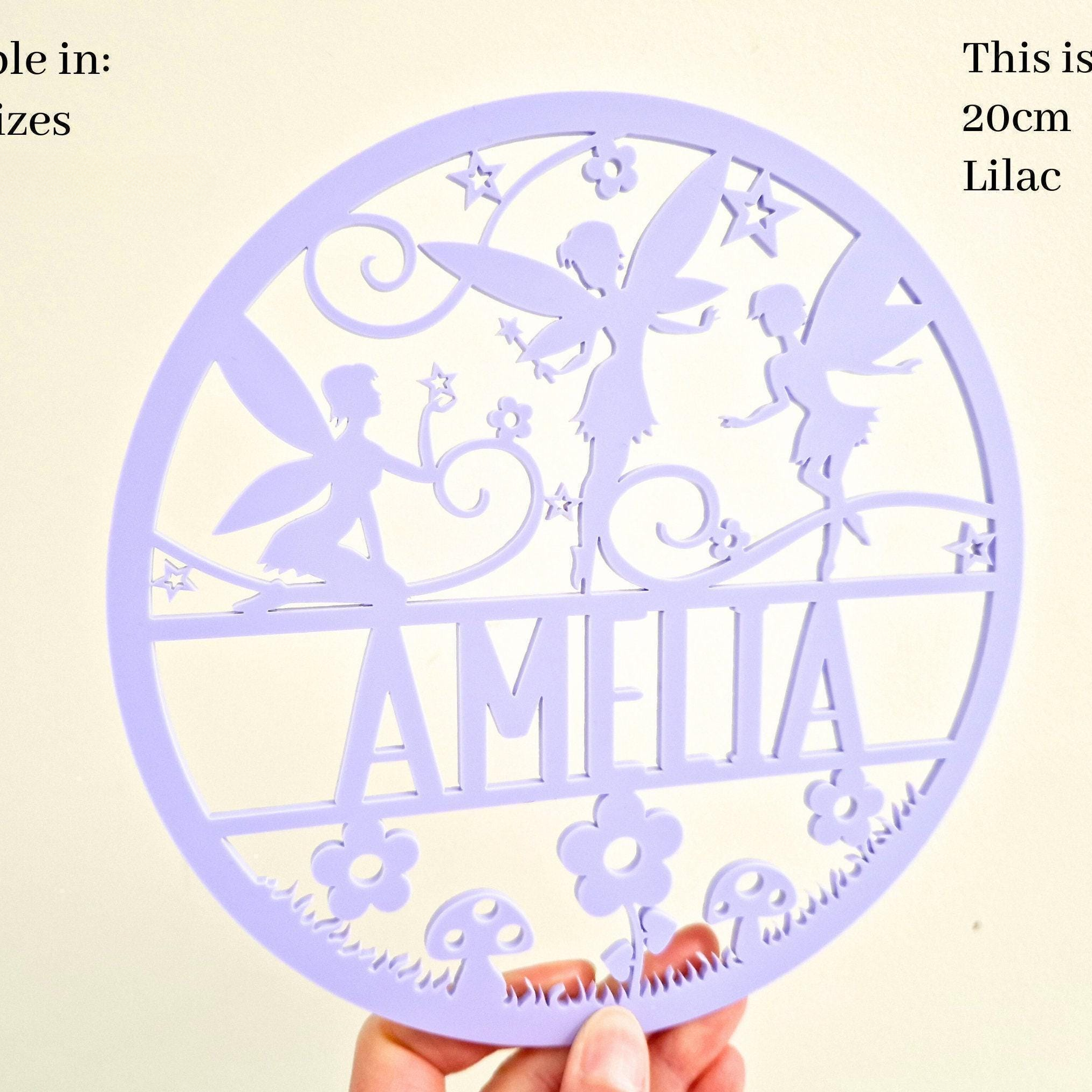 a hand holding a circular purple sign with a design featuring a fairy and flowers. The sign has the name "Amalia" written on it.