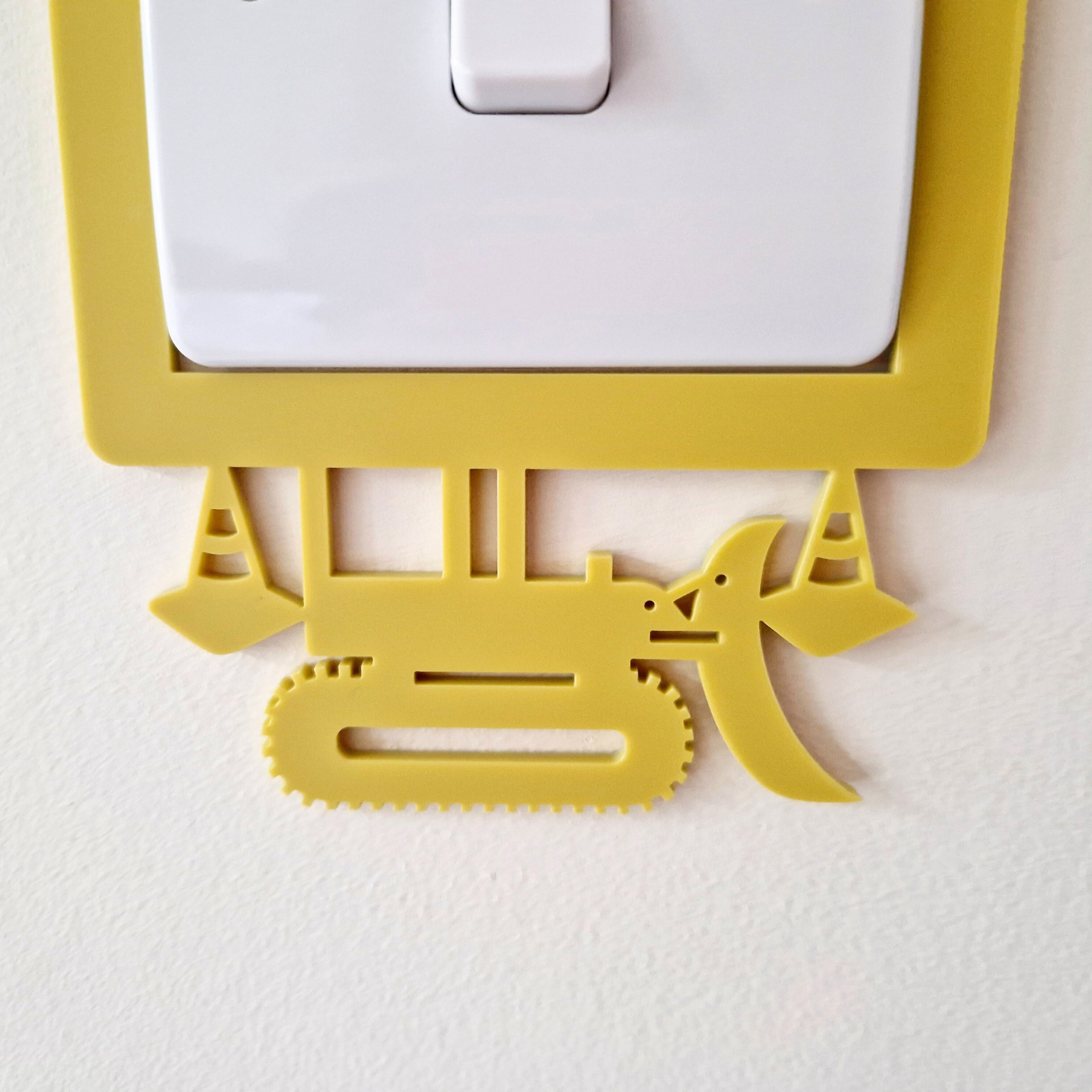A yellow plastic construction vehicle-shaped light switch cover is attached to a white wall.