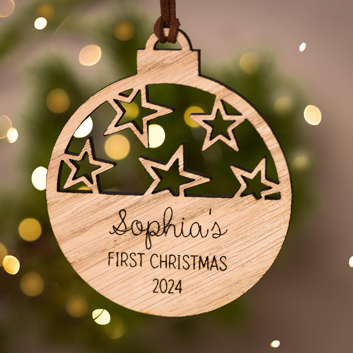 Wooden ornament with cut-out stars and engraved text 'Sophia's First Christmas' on a blurred Christmas tree background