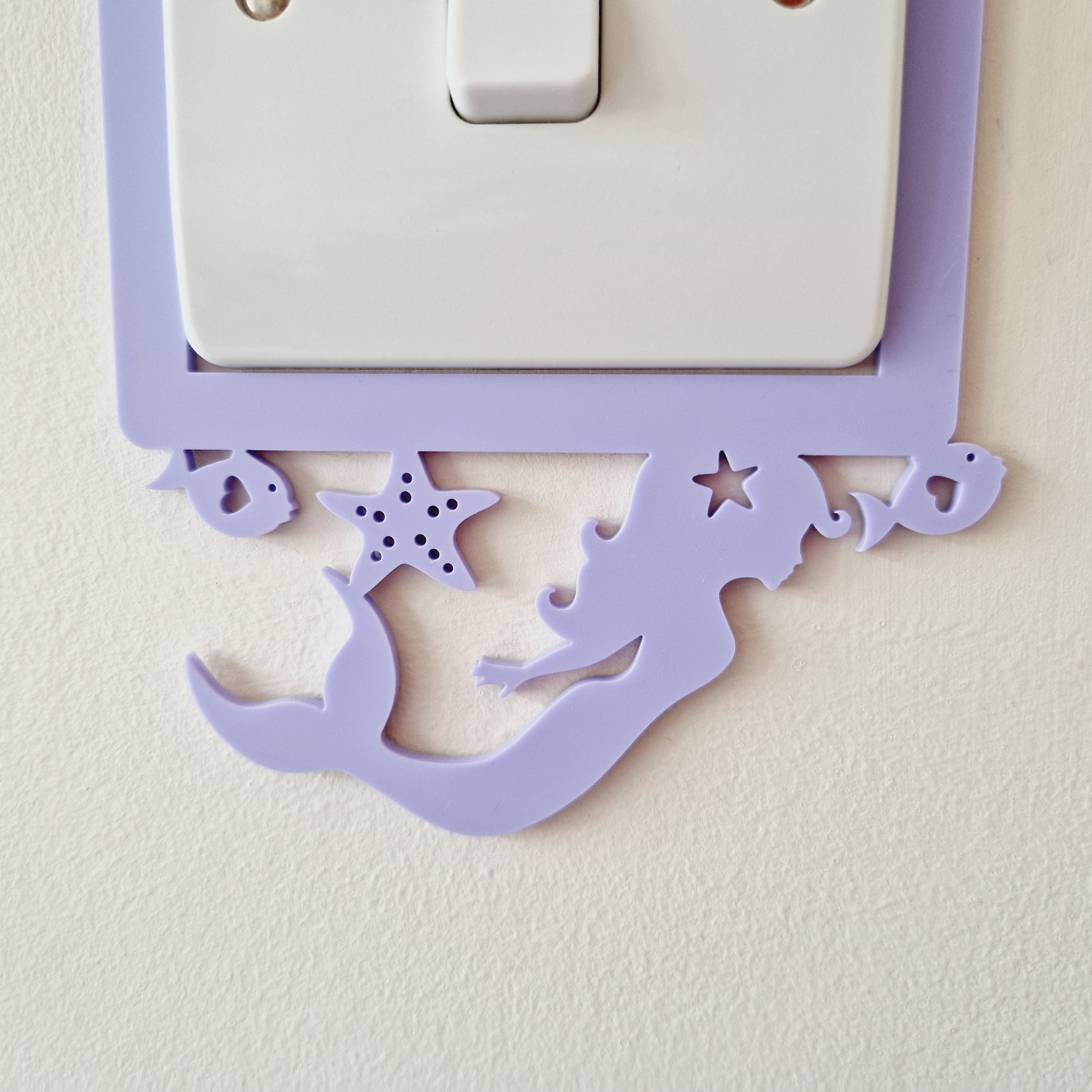 A purple mermaid-shaped light switch cover is attached to a wall, with a white light switch below it.