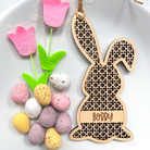 a wooden Easter bunny decoration hanging on a wall, surrounded by colorful Easter eggs and pink felt flowers.