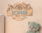 A wooden nameplate with the name "Sophie" is hanging on a wall, surrounded by various sea animal silhouettes. Next to the nameplate is a stuffed teddy bear.