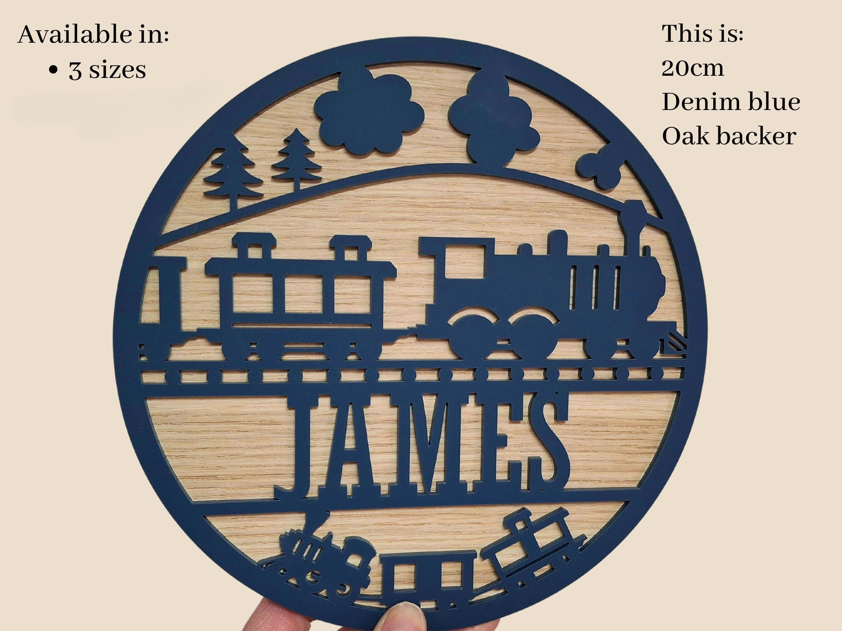 A hand holding a circular wooden sign with a train design and the name "JAMES" written on it.
