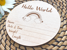 A wooden sign with the text "Hello World" and a rainbow graphic, placed on a woven placemat with a white daisy flower.