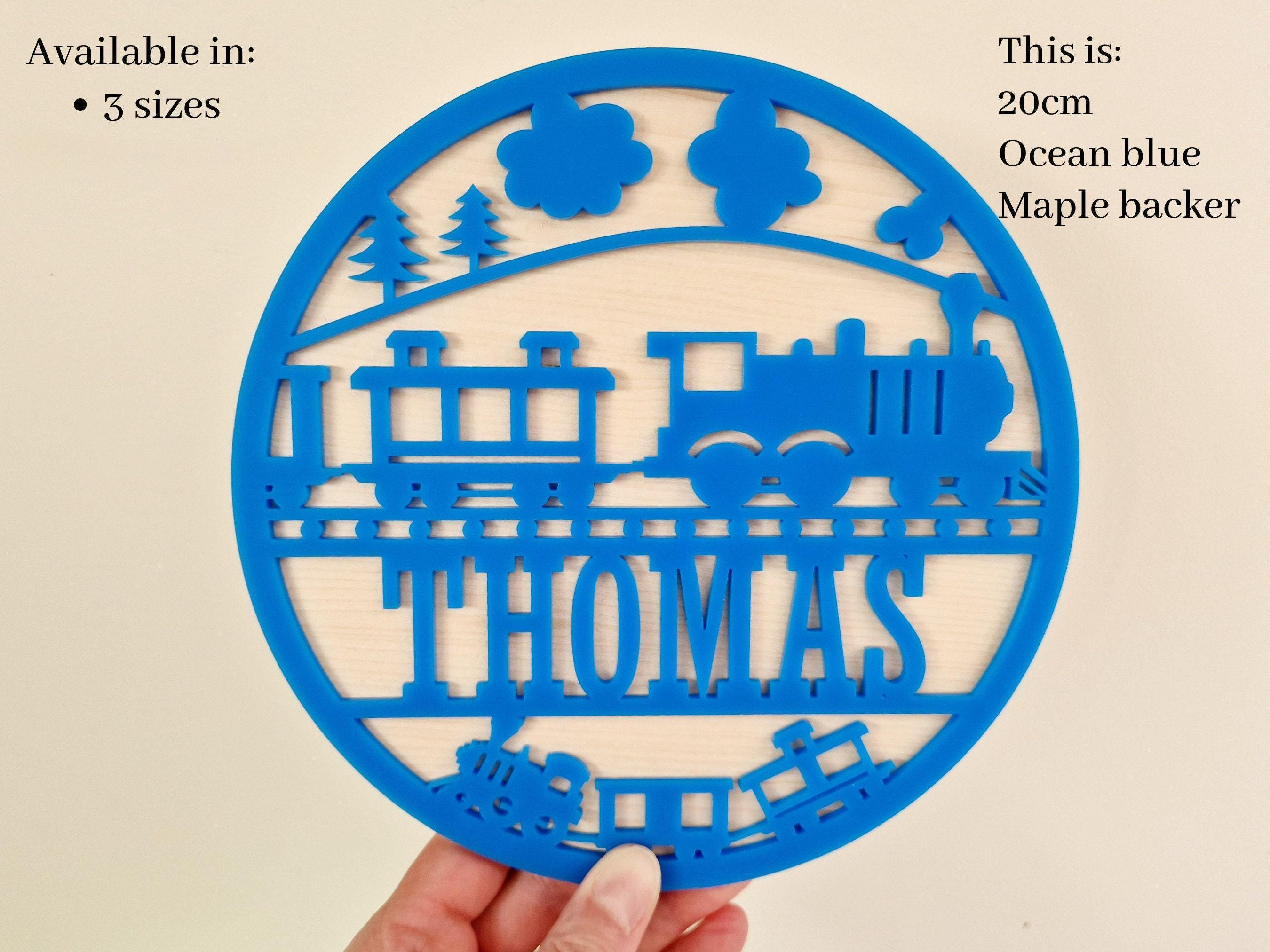 A hand holding a blue circular cutout with a train design and the name "THOMAS" written on it.
