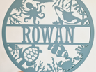 a round sign with the name "Rowan" on it, available in 25+ colors and 3 sizes. The sign is described as a thoughtful gift for various occasions, such as birthdays, Christmas, baby showers, and other special events. It is also wrapped in tissue paper, ready for gifting.
