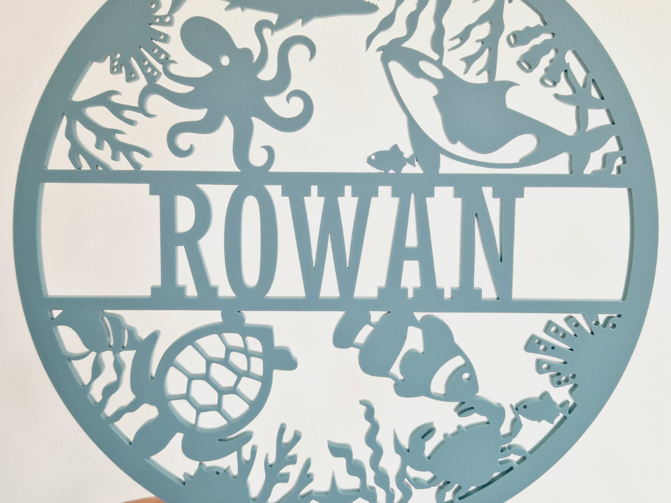 a round sign with the name "Rowan" on it, available in 25+ colors and 3 sizes. The sign is described as a thoughtful gift for various occasions, such as birthdays, Christmas, baby showers, and other special events. It is also wrapped in tissue paper, ready for gifting.