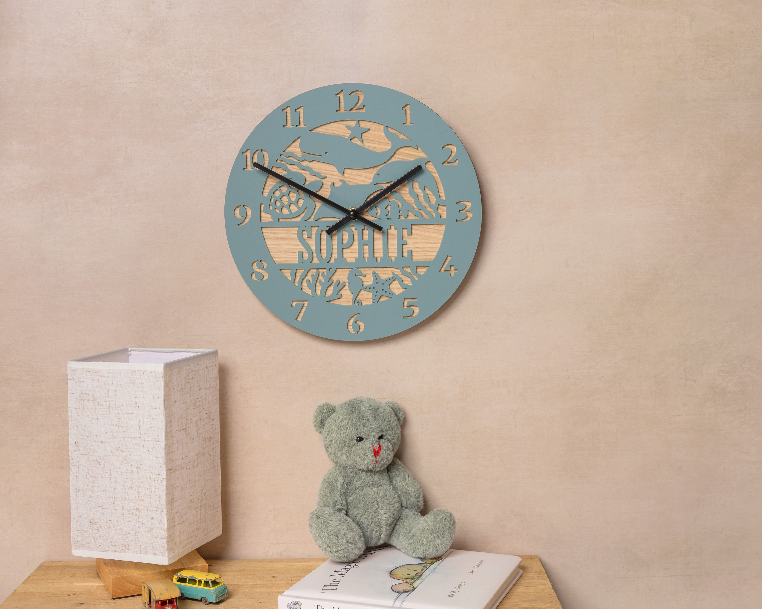 a wall clock with a personalized name, a teddy bear, and a lamp on a wooden table.