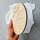A wooden birth announcement plaque is being held up, with a white baby onesie visible in the background.