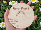 A wooden postcard with a rainbow graphic and the text "Hello World" is being held in front of a background of white daisies.