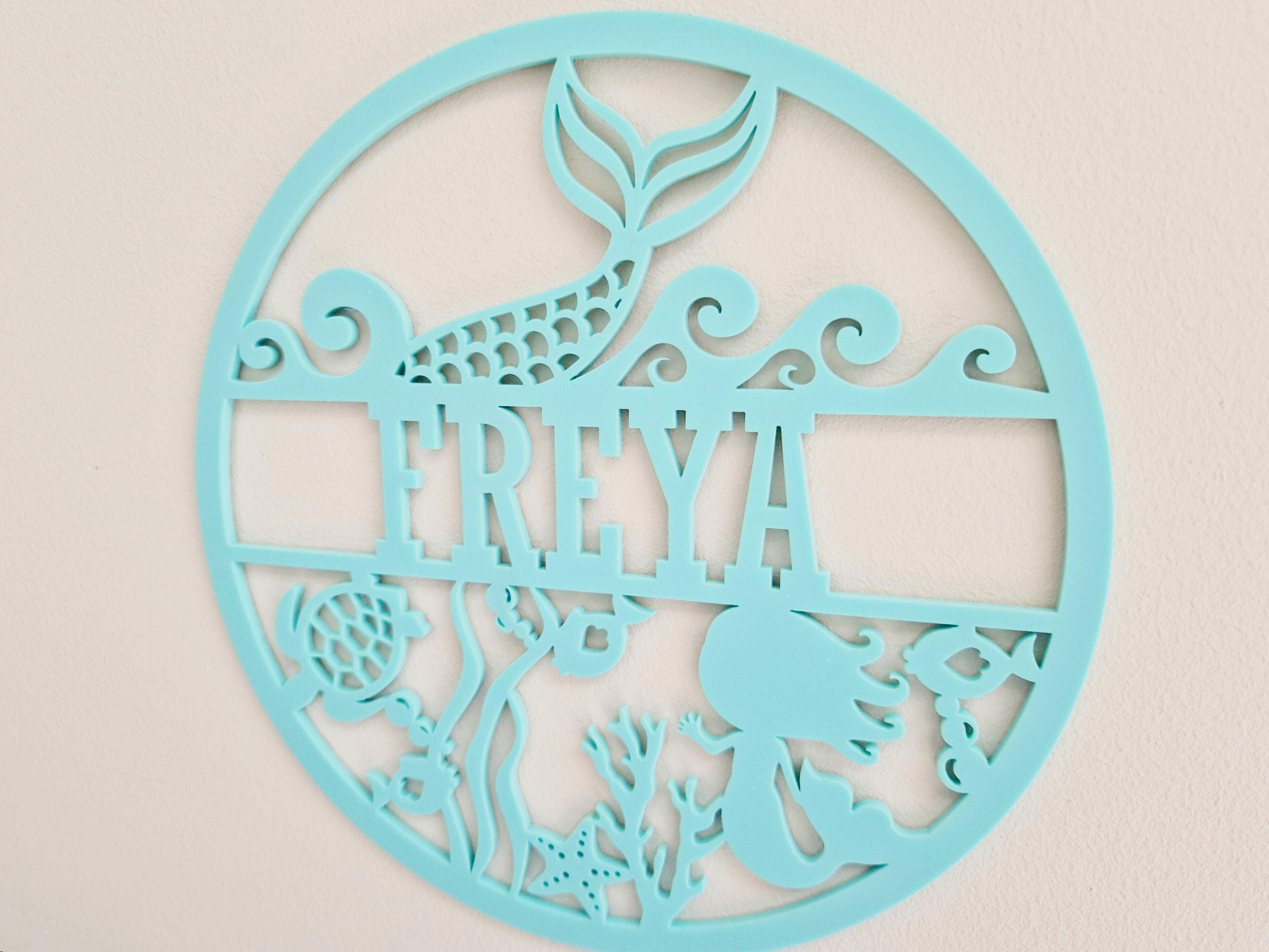 A round, light blue wall hanging featuring a mermaid silhouette and various marine-themed elements, including a fish, sea star, and coral.