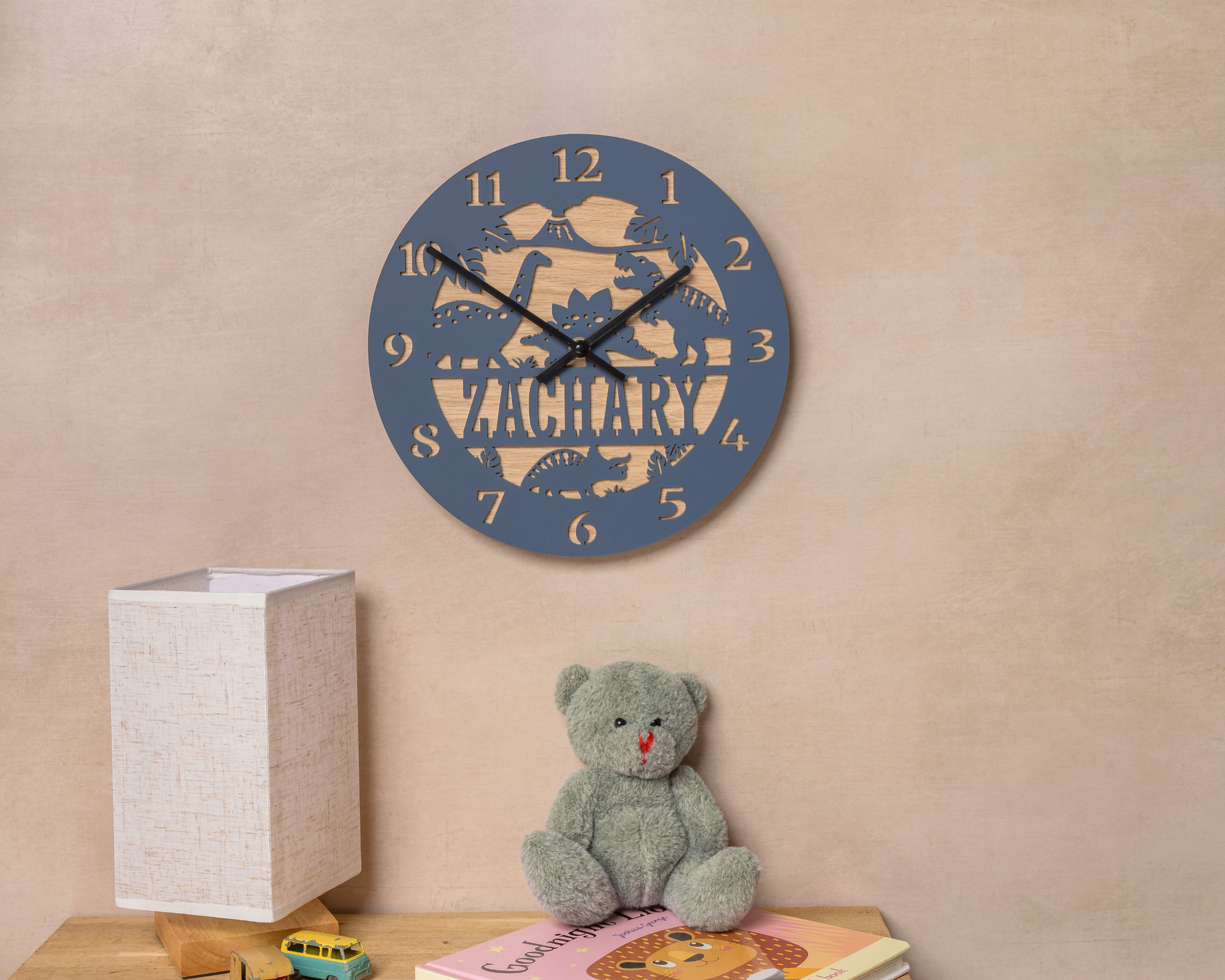 A wall clock with the name "ZACHARY" printed on it, along with a teddy bear and a children's book on a shelf.