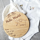 A wooden baby announcement card with a name and date and time information, along with a baby's weight and length, is placed next to a white onesie with animal prints.