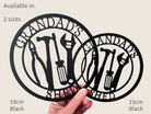 A person is holding two metal cutouts of a design featuring tools and the text "GRANDDAD'S SHED".