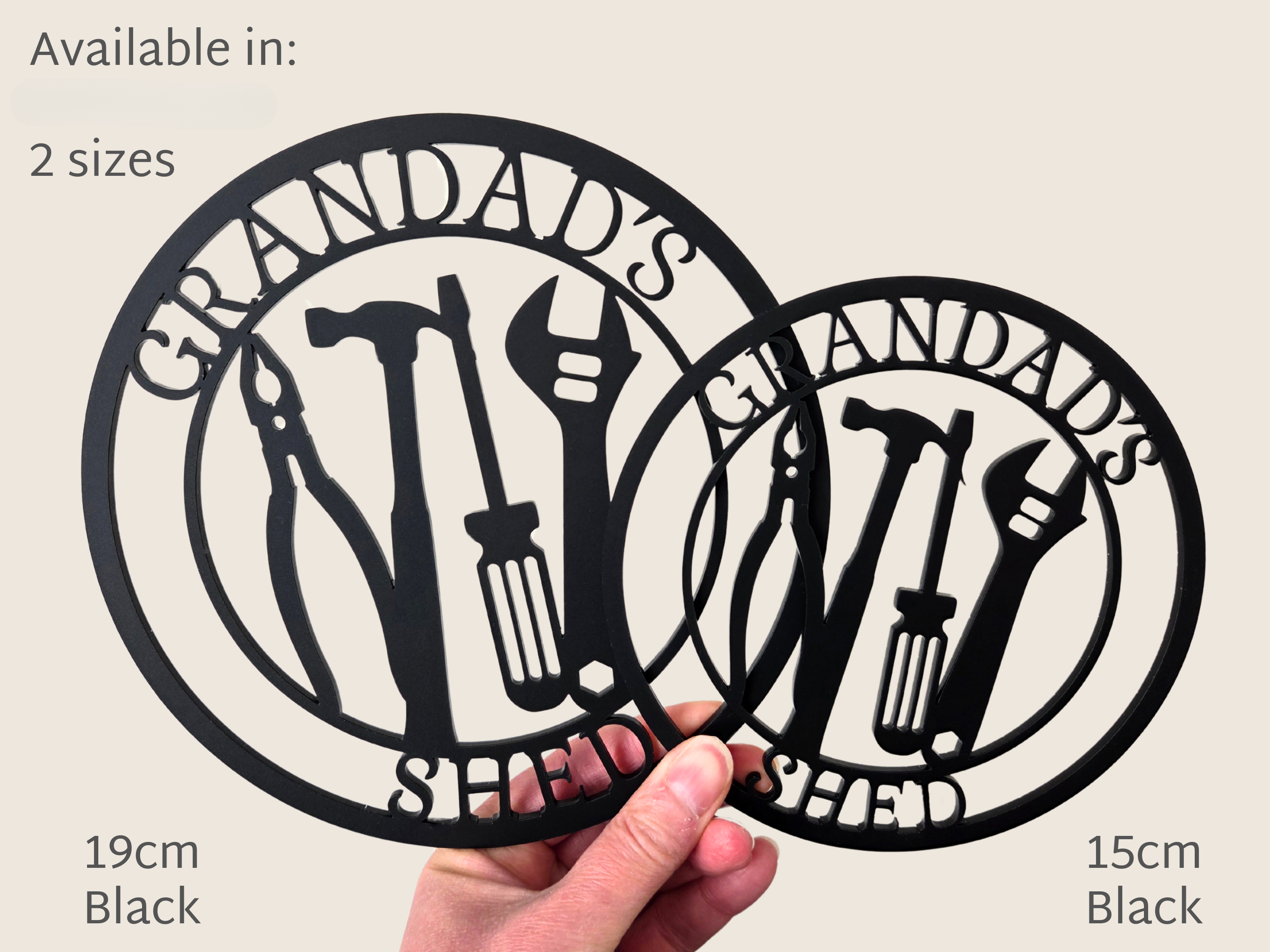 A person is holding two metal cutouts of a design featuring tools and the text "GRANDDAD'S SHED".