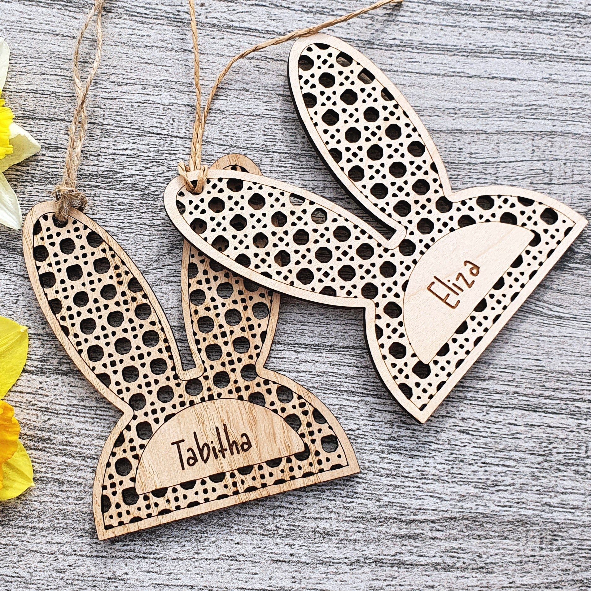 Wooden bunny ear-shaped tags with engraved names next to yellow daffodils on a wooden surface