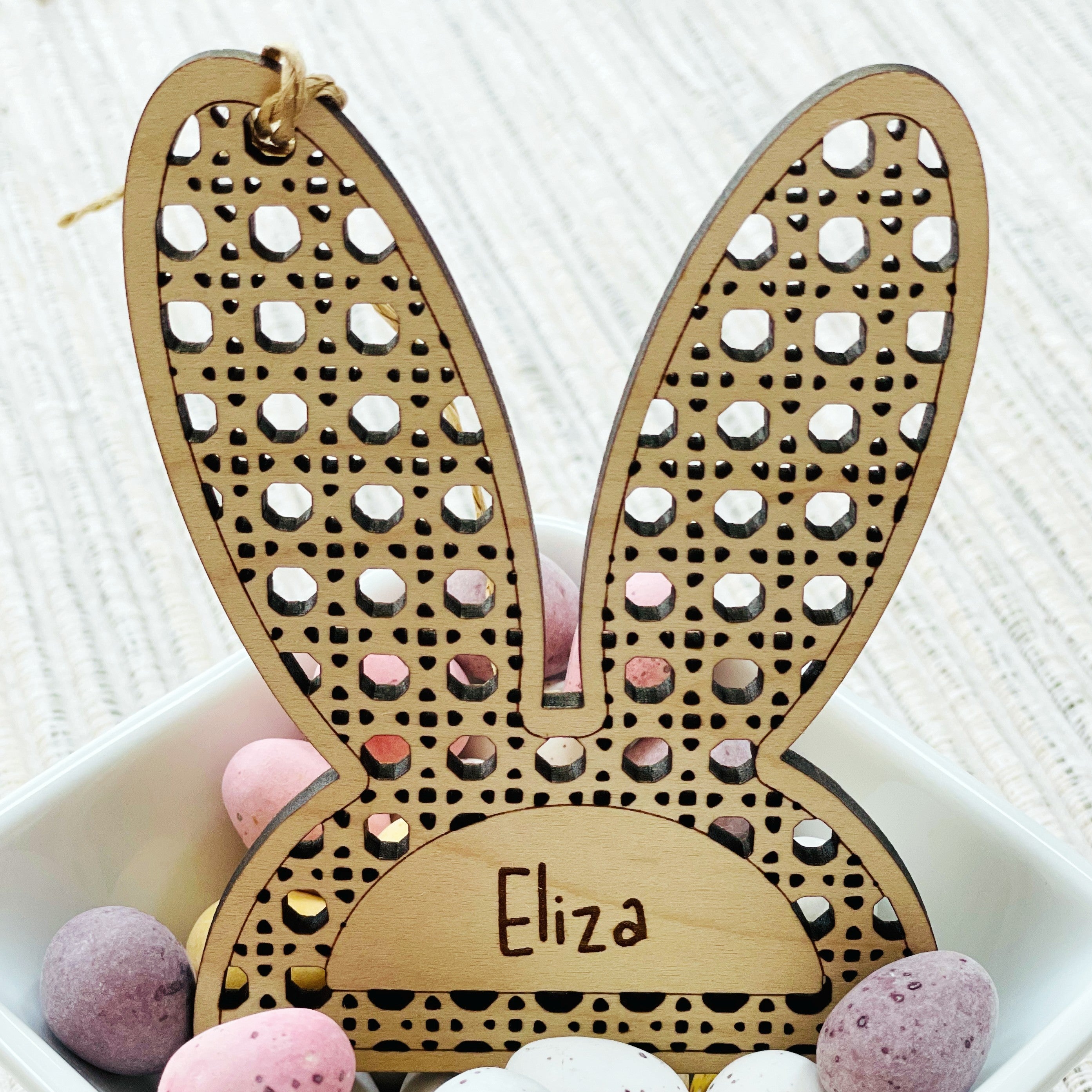 Bunny-shaped ears ornament with 'Eliza' engraved, sat in a bowl filled with colorful Easter eggs on a white surface.