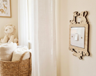 A light switch cover featuring a natural wood frame with animal cutouts of a giraffe, elephant, and monkey, mounted on a beige wall next to a window with sheer curtains.