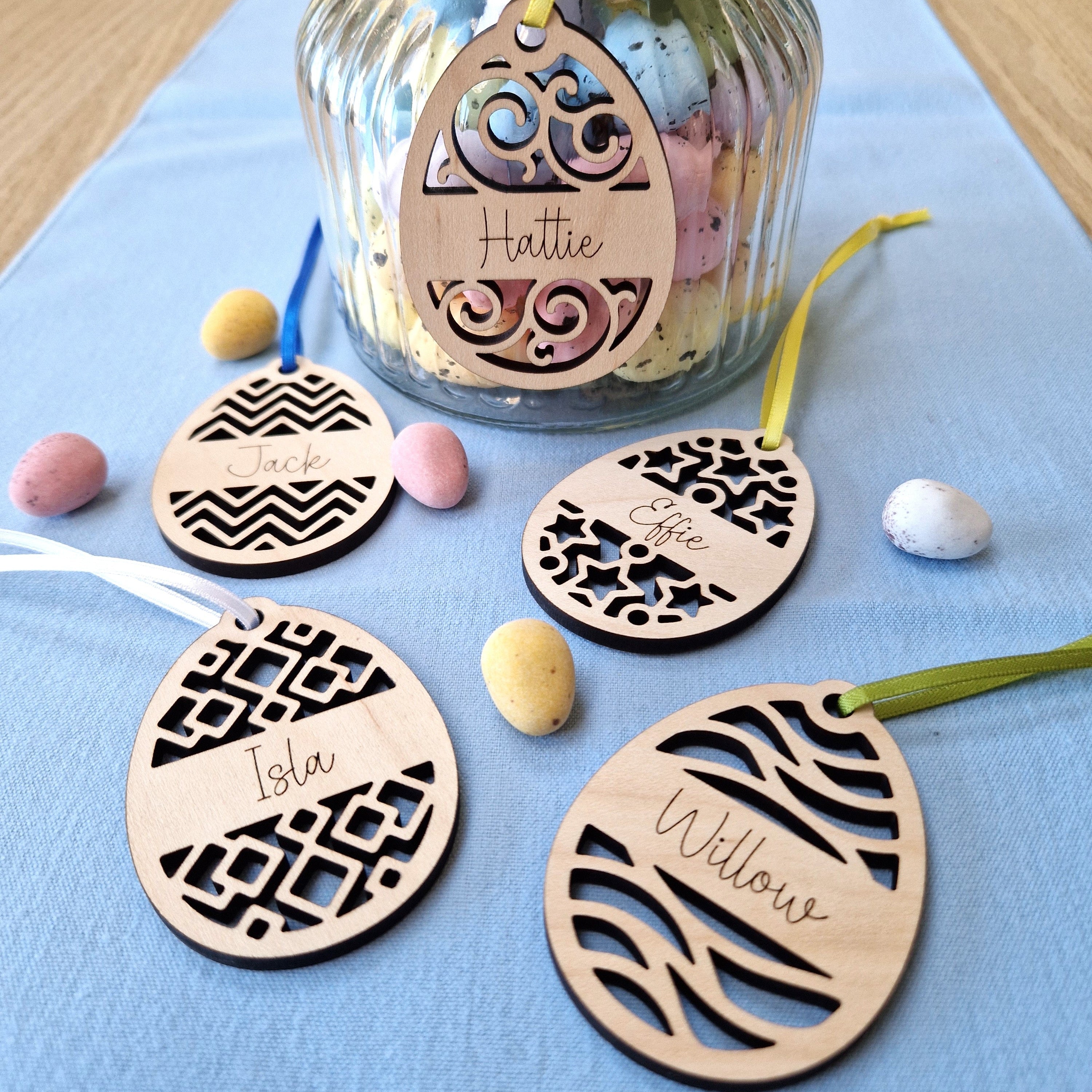 Wooden easter egg name tags with decorative geometric patterns on a blue surface with a jar of small eggs.