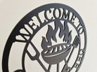 Close up of decorative acrylic sign with 'Welcome to Grandad's BBQ' text and grill icon on a beige background
