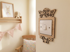 Decorative wooden butterfly light switch plate on a wall with a shelf and chair in the background.