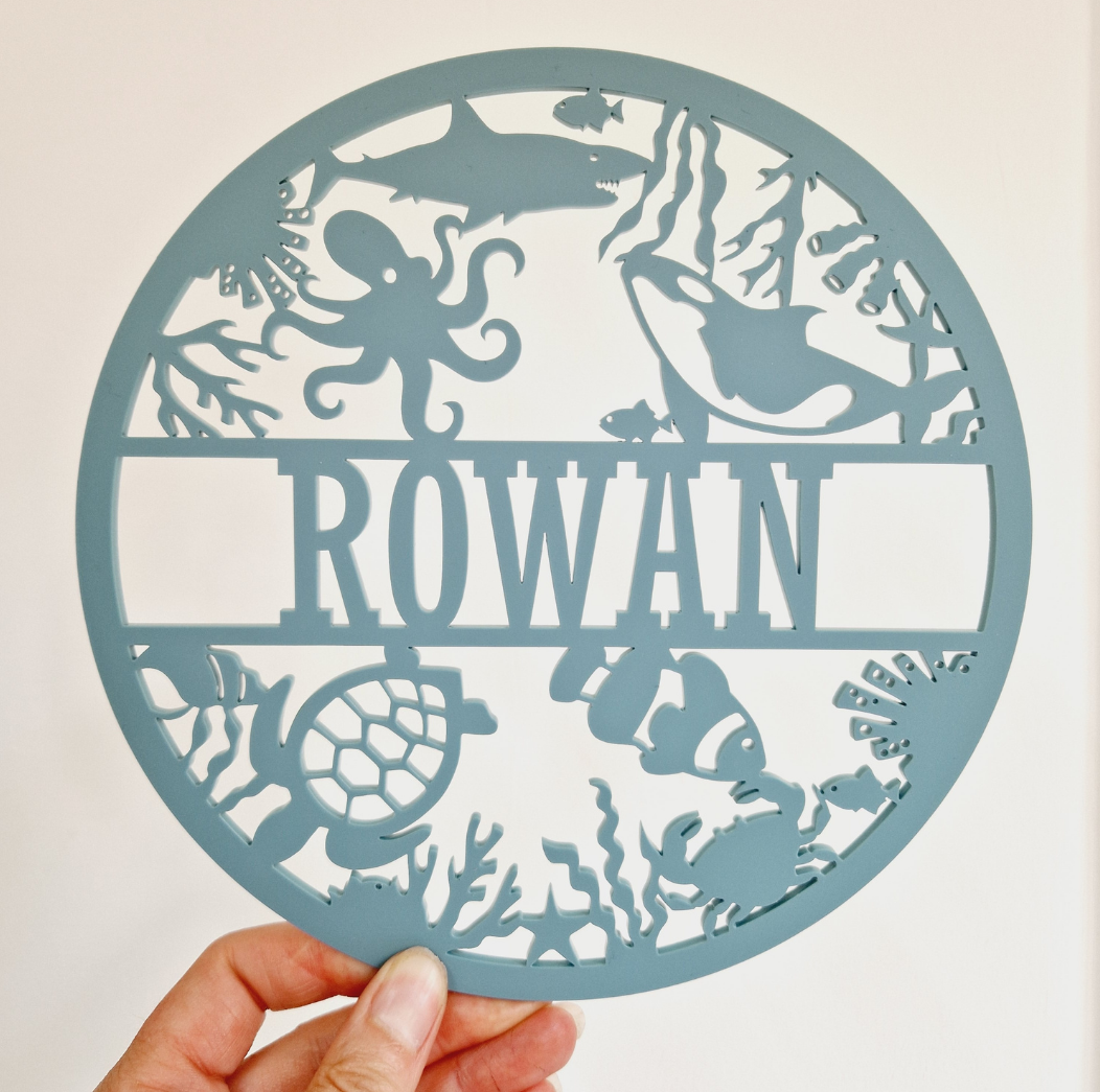 Round OCEAN theme name sign with 'ROWAN' text, held by a hand, on a light background with product details.