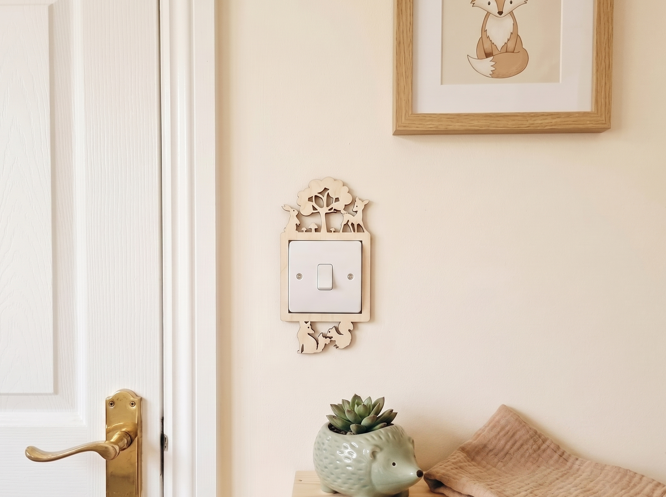 Decorative wooden wall switch plate with woodland forest animal design and underneath, a succulent plant and folded blanket on a bench.