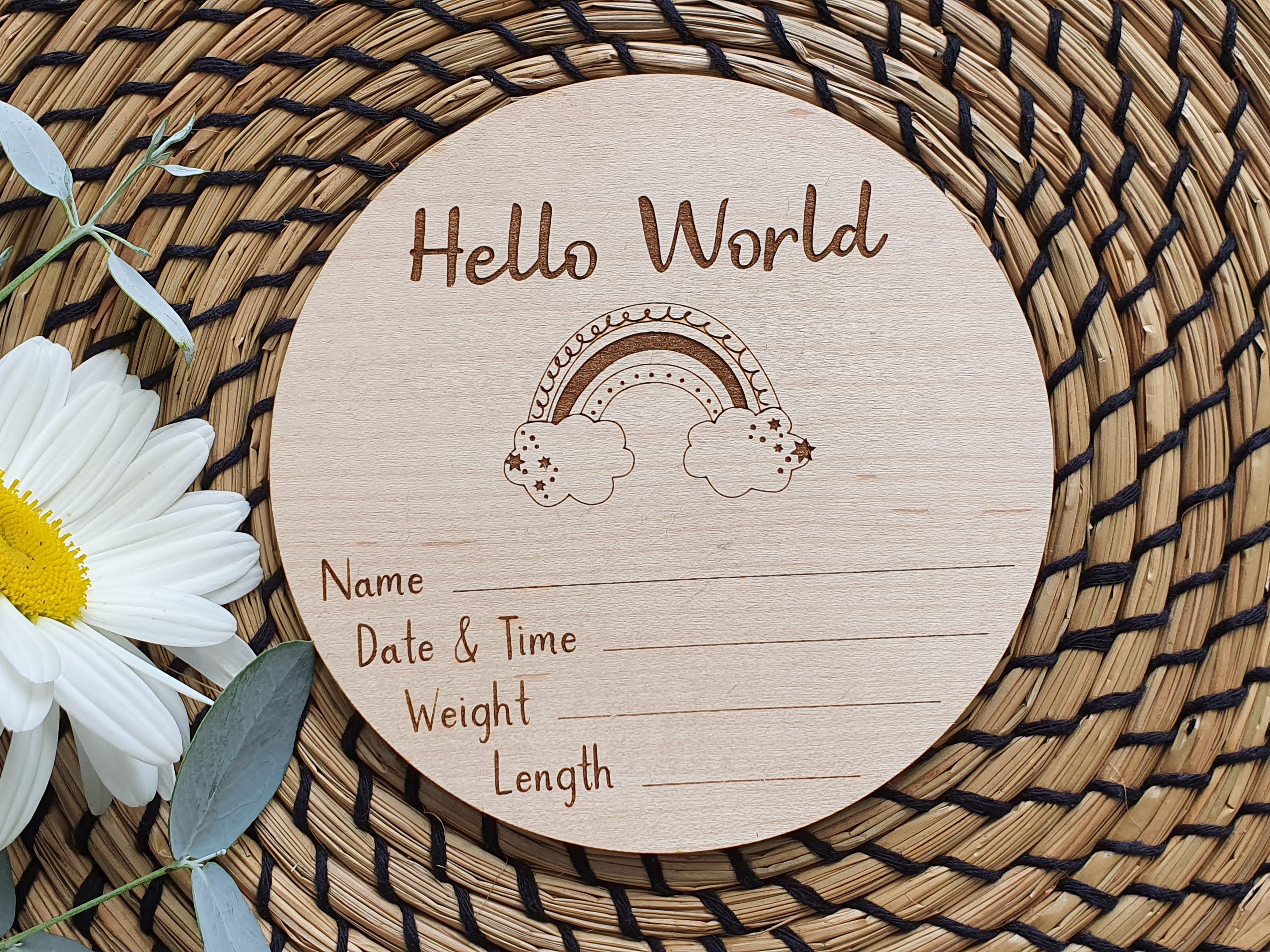 A wooden postcard with a rainbow graphic and the text "Hello World" is placed on a woven mat with a white daisy flower.