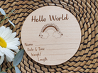 A wooden postcard with a rainbow graphic and the text "Hello World" is placed on a woven mat with a white daisy flower.