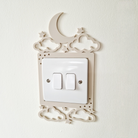 a white light switch with a decorative frame that includes a crescent moon, stars, and clouds.